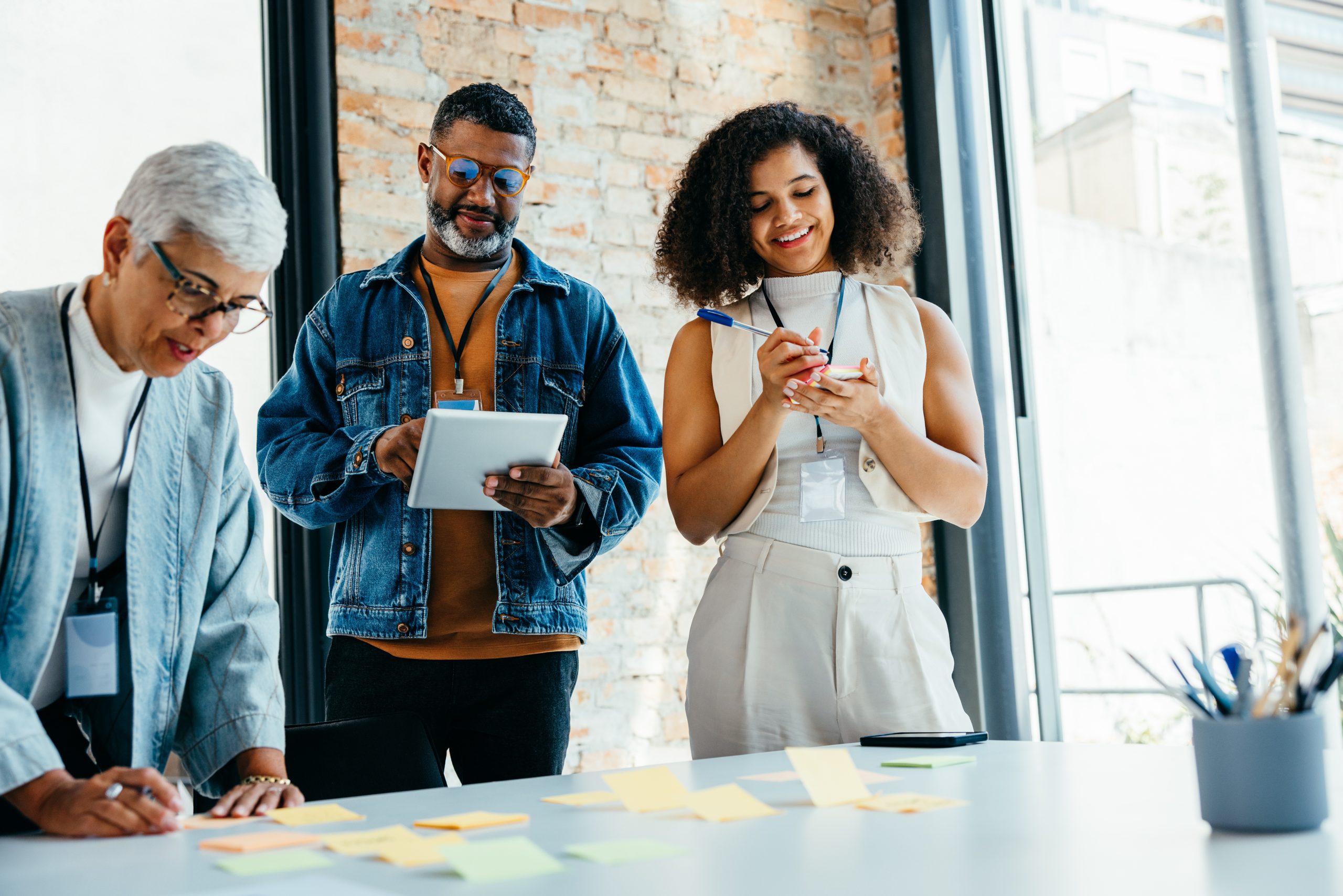 A multigenerational team of professionals collaborates in a well-lit modern office, brainstorming with sticky notes and digital tablets during a productive business meeting.