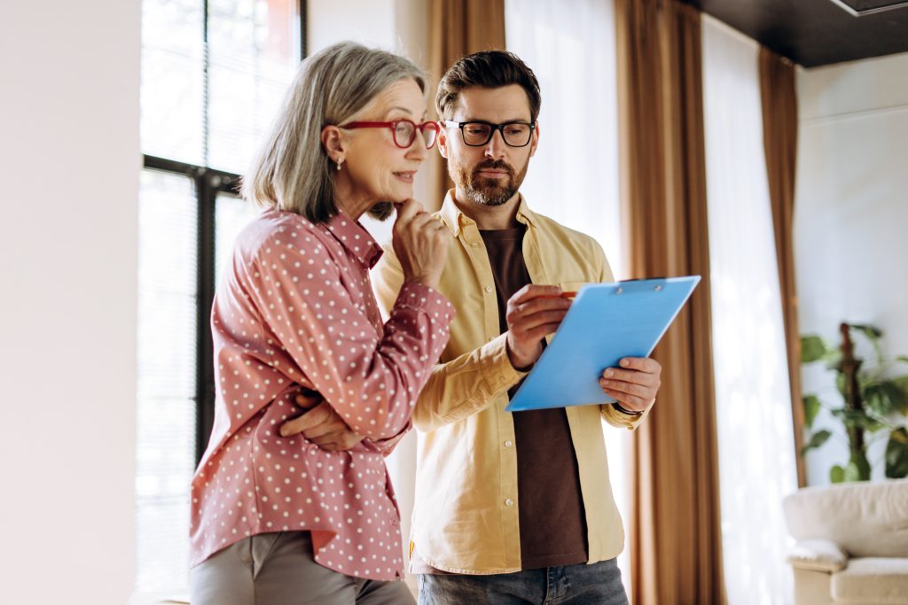Financial advisor holding a clipboard and discussing contract details with a thoughtful senior woman, carefully considering her investment options in a cozy living room setting