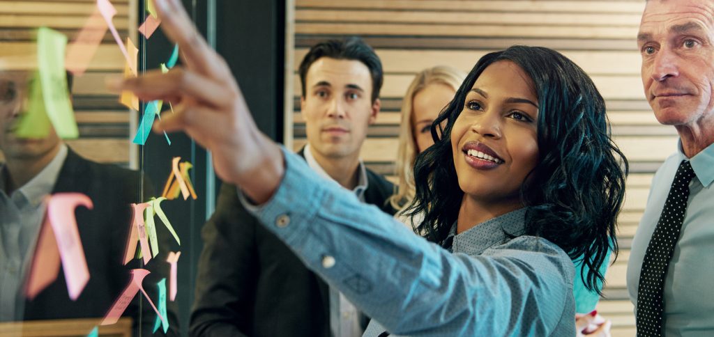 Young smiling black businesswoman at the wall with stickers communicating with her colleagues. Mixed team of entrepreneurs