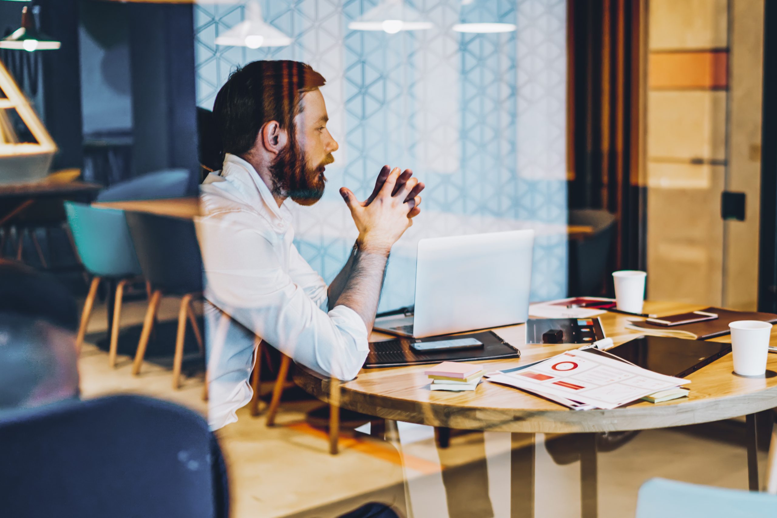 Thoughtful male entrepreneur dressed in formal wear thinking on development creative ideas for business project.Pondering proud ceo sitting at meeting table behind glass wall of modern office